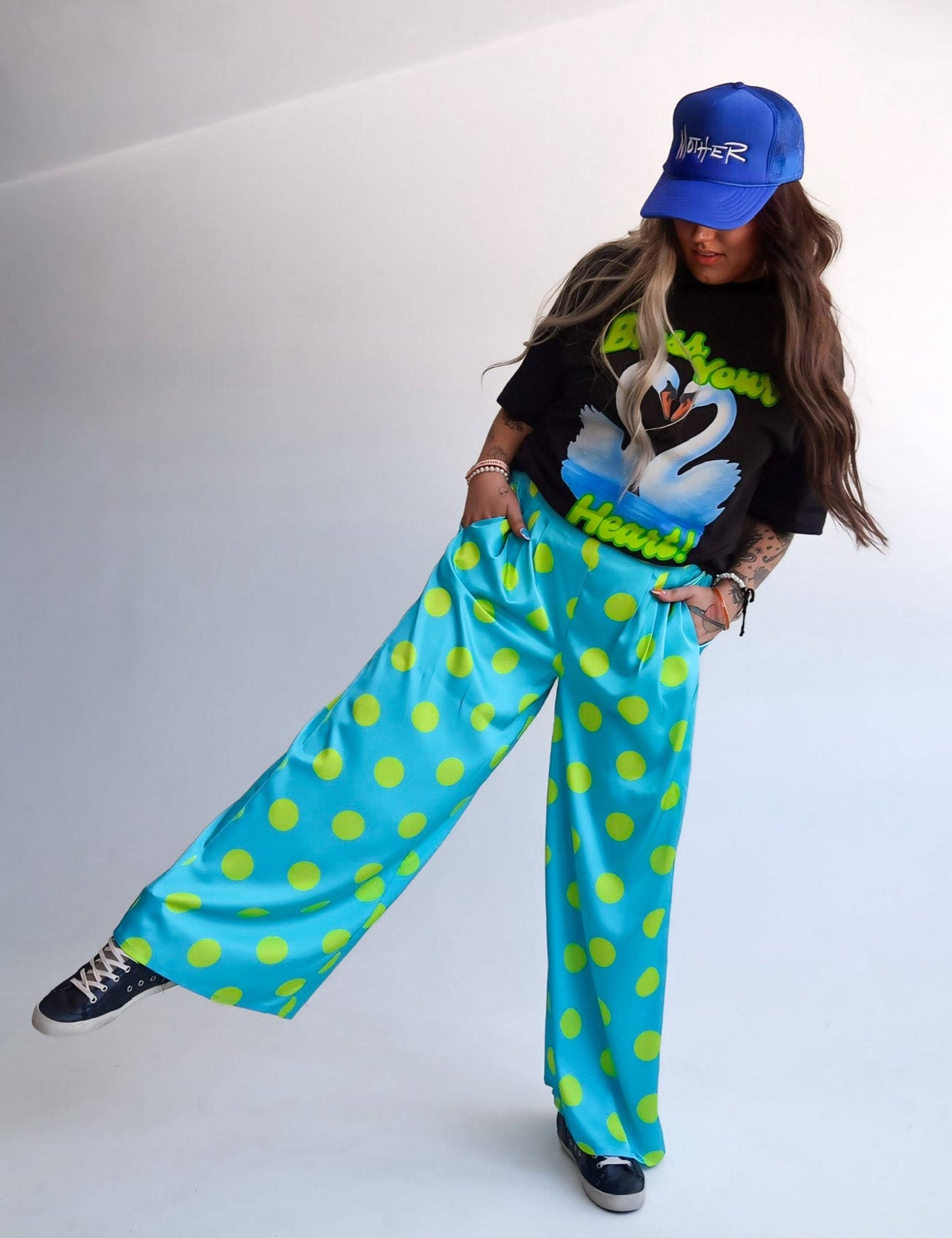 Person wearing a black t-shirt with a graphic design, blue cap, and bright blue pants with green polka dots on a white background.